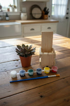 A miniature painting kit on a wooden tabletop, featuring a small wooden crate made from pallet wood, a small potted succulent, a paint palette with six colors, and a paintbrush.
