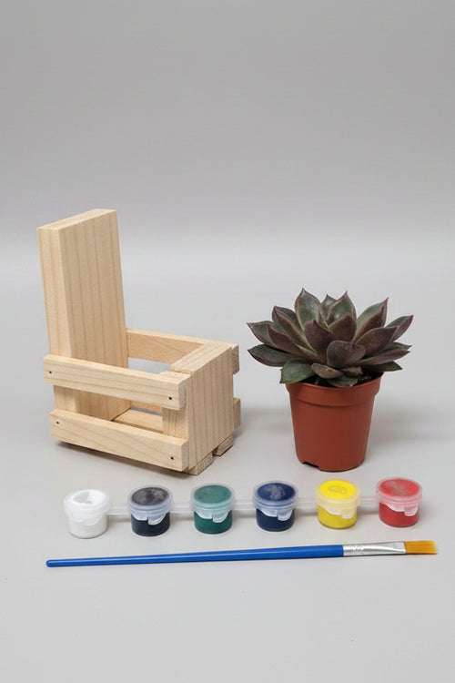 A miniature wooden painting set displayed on a wooden tabletop against a light gray wall. The set includes a small wooden crate made from pallet wood, a potted succulent, five miniature paint pots in white, gray, green, yellow, and red, and a small blue-handled paintbrush.