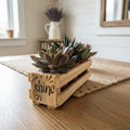 A small rectangular wooden planter box made from pallet wood with the word 'shine' printed on the side, holding a cluster of green succulent plants, set on a light wood tabletop against a neutral wall.